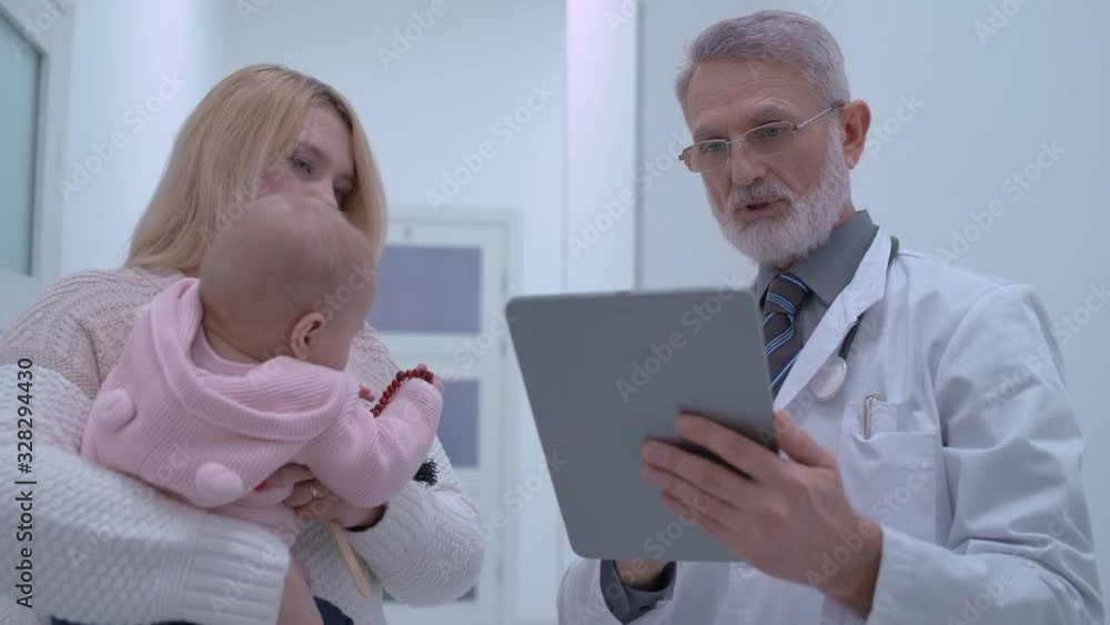 Male physician making appointment on tablet, consulting young mother with baby Male physician making appointment on tablet, consulting young mother with baby
