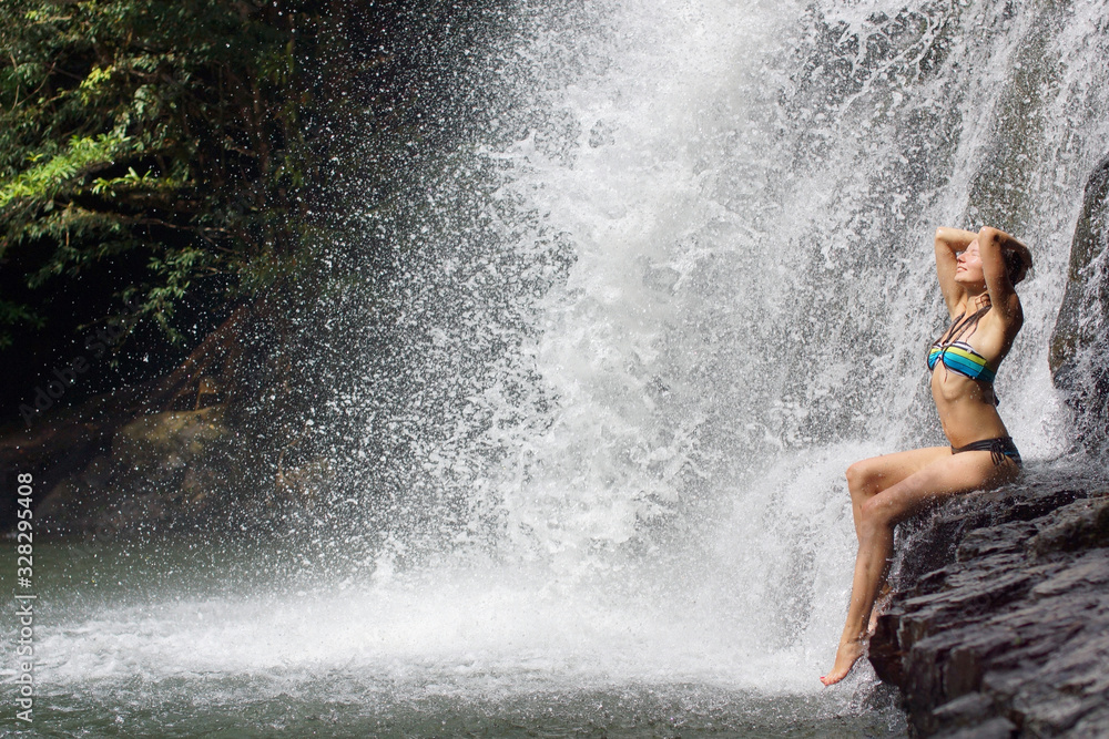 Young athletic woman with long hair sitting on stone on background of waterfall with many drops and tropical forest Young athletic woman with long hair sitting on stone on background of waterfall with many drops and tropical forest