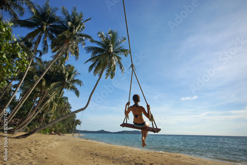 Young woman in bikini on rope swing on tropical sandy beach. Background of palm trees, blue sky and sea. View from back © Vladimir