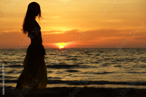 Black silhouette of woman with long hair, in long skirt standing on the beach during sunset and looking at sea