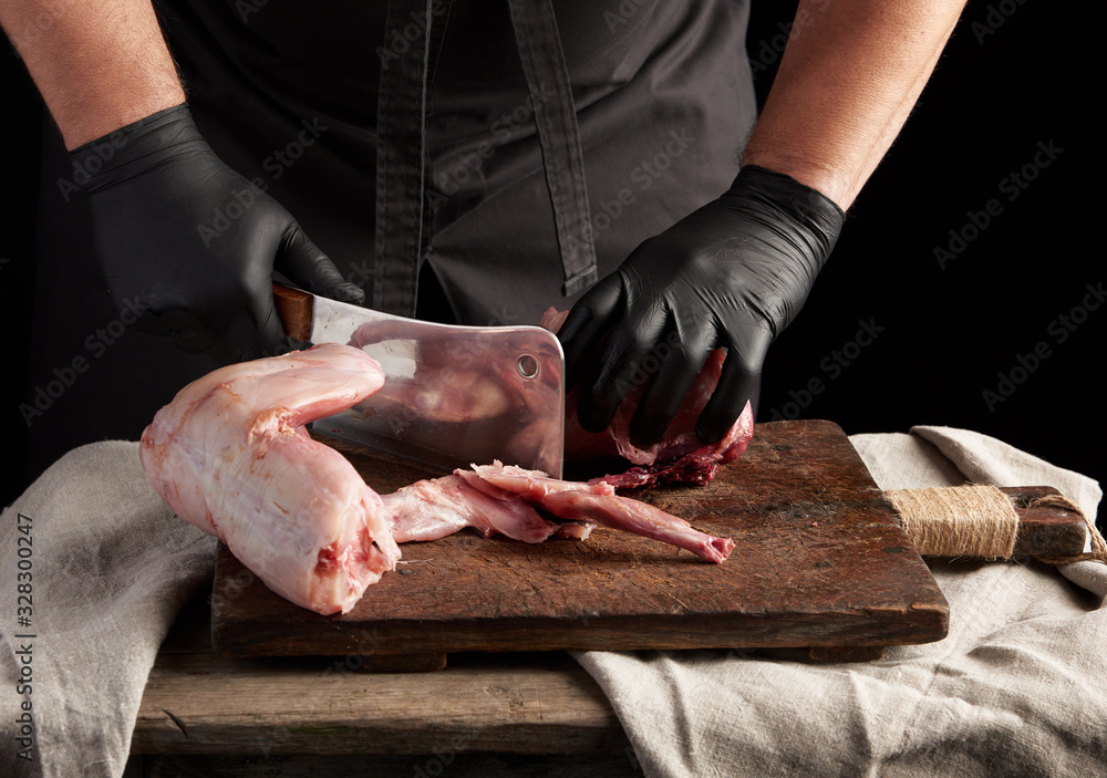 Chef in black latex gloves holds a big knife and cuts into pieces raw rabbit meat