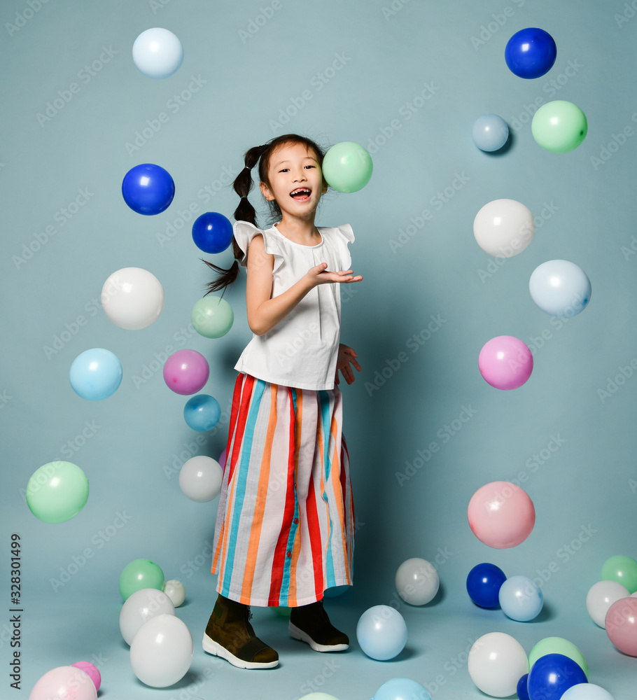 Fototapeta premium Laughing asian kid girl in party clothes is having fun surrounded by colorful air balloons holds hand up with open palm