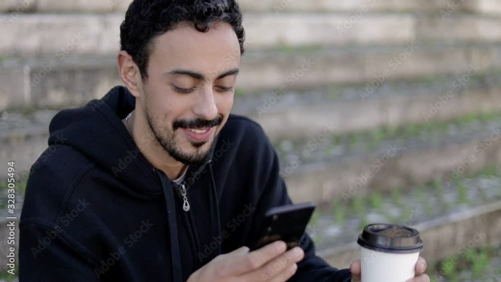 Content man with smartphone and coffee to go. Handsome happy young man sitting on steps drinking from paper cup and using mobile phone. Connection concept Content man with smartphone and coffee to go. Handsome happy young man sitting on steps drinking from paper cup and using mobile phone. Connection concept