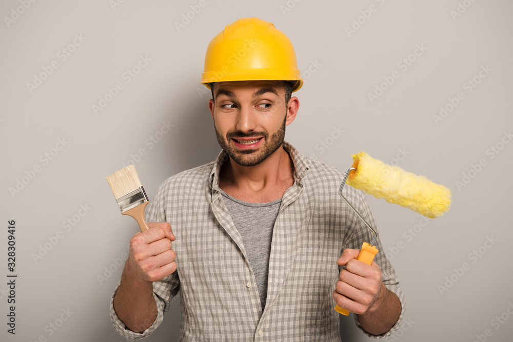 thoughtful manual worker in hardhat looking at paint roller and brush on grey thoughtful manual worker in hardhat looking at paint roller and brush on grey