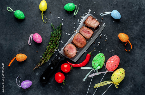 Grilled beef steak over a meat knife with spices and decorative Easter eggs. View from above. on a stone background. Easter celebration concept