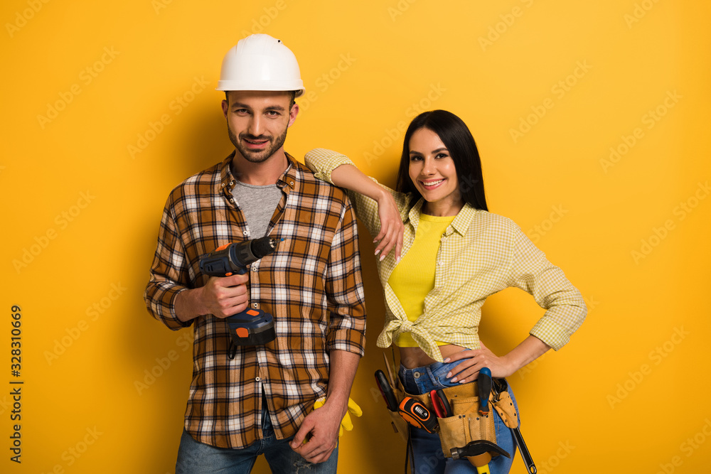 professional smiling manual workers with tool belt holding electric drill on yellow - obrazy, fototapety, plakaty 