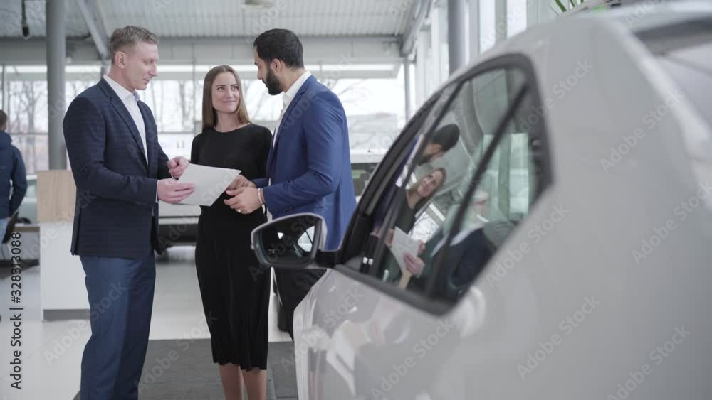 Adult successful family talking with car dealer in showroom. Young confident Middle Eastern husband and beautiful Caucasian wife choosing automobile in dealership. Wealth, luxury, business. Adult successful family talking with car dealer in showroom. Young confident Middle Eastern husband and beautiful Caucasian wife choosing automobile in dealership. Wealth, luxury, business.