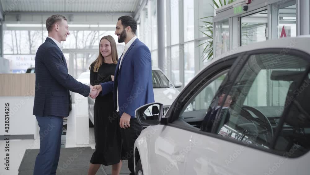 Young rich multiracial family buying car in dealership. Smiling Middle Eastern client and Caucasian dealer shaking hands in showroom. Agreement, business, luxury, success. Young rich multiracial family buying car in dealership. Smiling Middle Eastern client and Caucasian dealer shaking hands in showroom. Agreement, business, luxury, success.