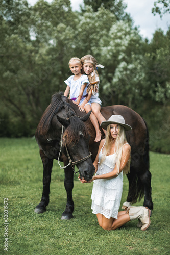 Happy family, mother with two children cute daughters riding a horse. Mother in white dress, hat and cowboy boots standing on the knees and feeding the horse. Family time on nature, horse riding