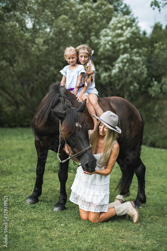 Attractive family, mother and two daughters wearing stylish casual boho clothes, walking outdoors on the meadow on a brown horse. Pretty young mother petting horse and two kids riding © sofiko14