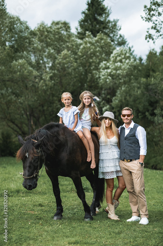 Stylish young ranch family, father, mother and two girls daughters riding a horse in a park or forest on a sunny summer day. Happy family in boho cowboy wear with horse outdoors