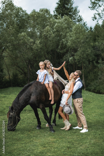 Loving family, father, mother and two little daughters riding horse at countryside outdoors. Young happy family having fun, giving high five to each other, family time concept. Summertime.