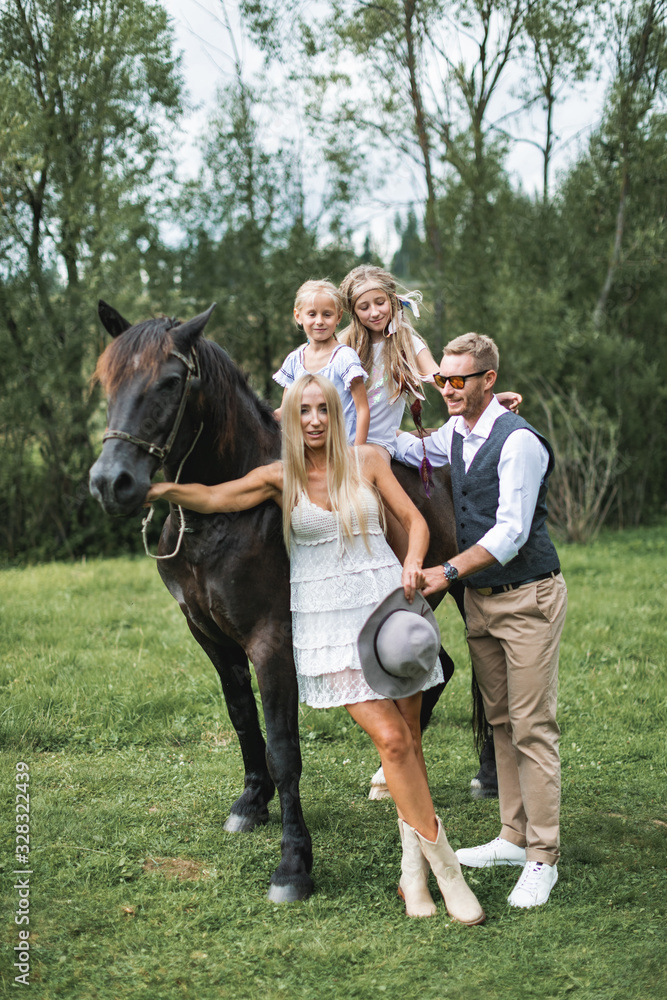 Cute two girls sisters riding horse on nature background, having walk with their mother and father in stylish boho cowboy wear, looking at camera and smiling. Stylish young family walking with horse