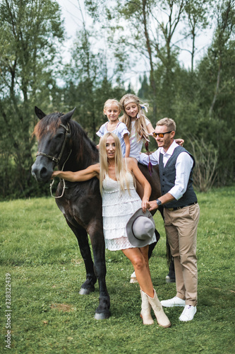 Cute two girls sisters riding horse on nature background, having walk with their mother and father in stylish boho cowboy wear, looking at camera and smiling. Stylish young family walking with horse © sofiko14