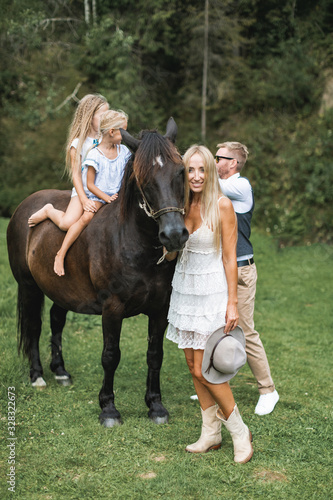 Couple, husband and wife, petting brown horse with pleasure in shiny sunlight in summer. Two little girls sisters daughters sitting on the horse and smiling. Outdoors, countryside, ranch lifestyle © sofiko14