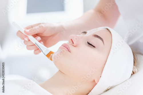 Close-up of hands of cosmetologist woman applying healing cream with brush on patient's face to young pretty caucasian woman lying on couch in beauty salon. Concept of antiaging facial skin treatments