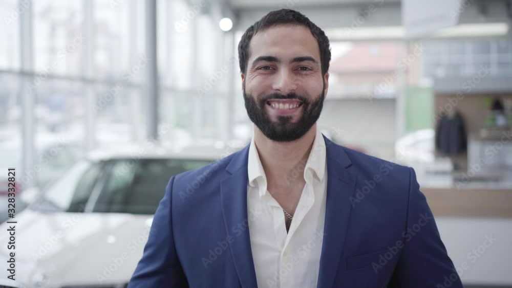 Portrait of successful Middle Eastern man posing in car dealership. Handsome young businessman looking back at new white vehicle, turning to camera and smiling. Success, lifestyle, business. Portrait of successful Middle Eastern man posing in car dealership. Handsome young businessman looking back at new white vehicle, turning to camera and smiling. Success, lifestyle, business.