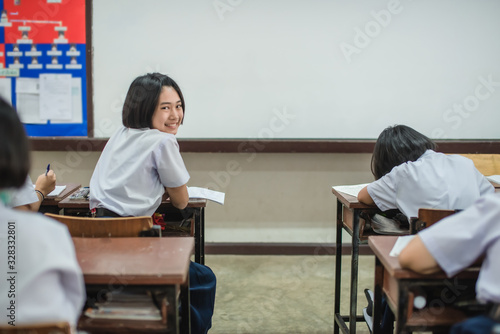 A pretty smiling Asian female student in white uniform is turning her face and smiling among her friends while studying in the classroom.
