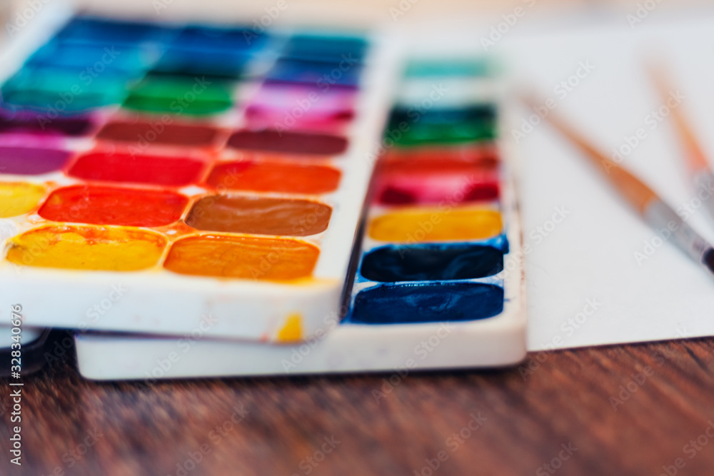 Watercolor paint, brushes well used and sheet of paper on wooden table with stack of books background. Watercolor paint, brushes well used and sheet of paper on wooden table with stack of books background.