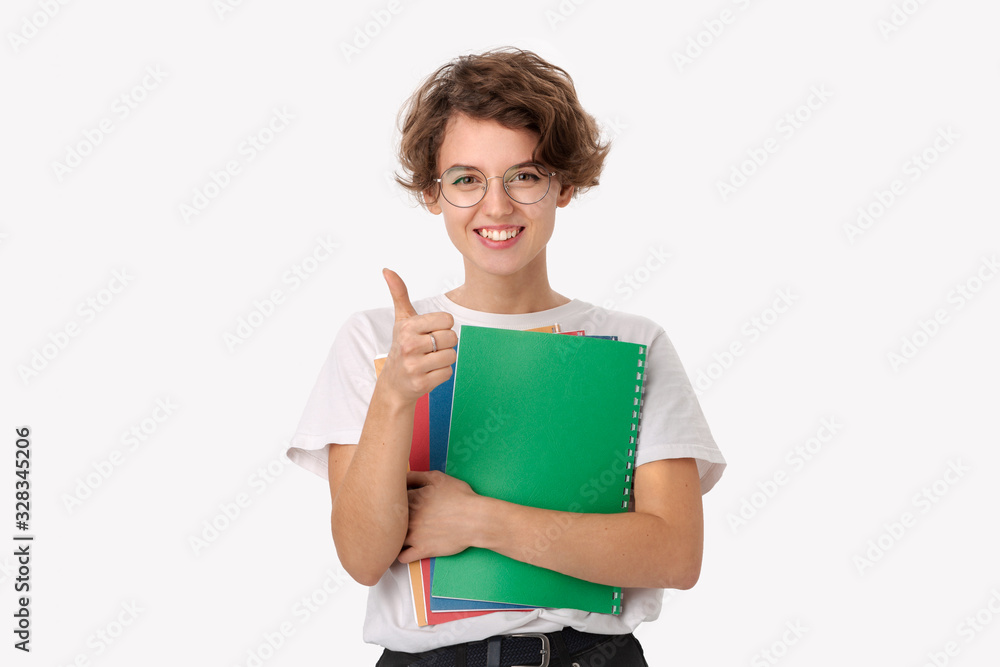 Excited young student in a white shirt and eyeglasses holding colorful folders and showing thumb up Excited young student in a white shirt and eyeglasses holding colorful folders and showing thumb up