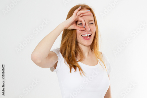 Red head girl in colorful clothing holding her fingers around the eye copy space isolated, young woman shows okays and looking through