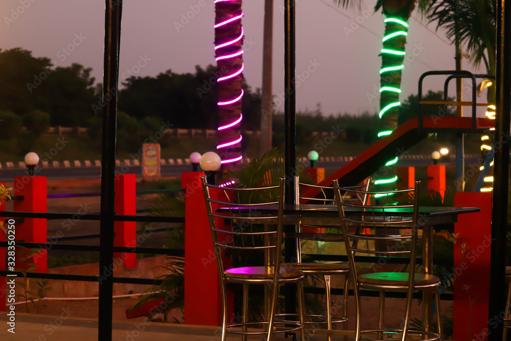 Cinematic View Of Exterior Garden Restaurant Bar With Table And Chairs Colorful Lights On Trees Late At Night