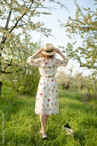 Rear view of a young woman with curly hair puts on a stylish wicker hat while walking in a green flowered garden. Spring mood © Kate