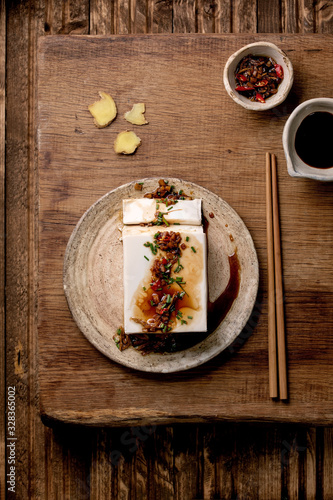 Silk tofu japanese soy cheese whole piece with chili ginger, chive and soy sauce topping on ceramic plate with chopsticks over wooden table. Flat lay, space