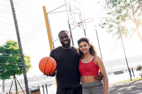 Outdoors Activity. African couple standing on basketball court with ball hugging smiling joyful