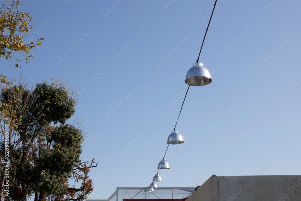 Round metallic lamps hanging between the square. Blue sky and trees in the background. Daylight