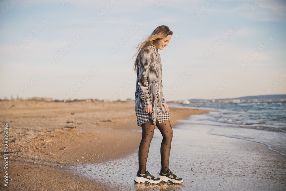 a pretty girl in a plaid dress and jacket walks along the sandy beach