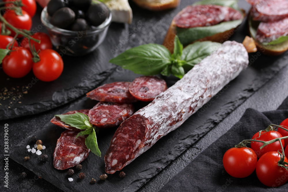 The concept of Italian cuisine. Sausage on a black board with tomatoes, olives, basil and cheese. Bread with sausage, sandwich. Background image, copy space The concept of Italian cuisine. Sausage on a black board with tomatoes, olives, basil and cheese. Bread with sausage, sandwich. Background image, copy space