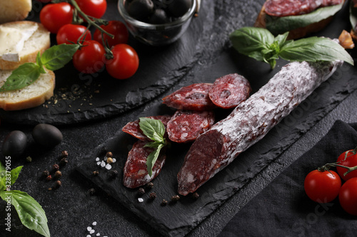 The concept of Italian cuisine. Sausage on a black board with tomatoes, olives, basil and cheese. Bread with sausage, sandwich. Background image, copy space