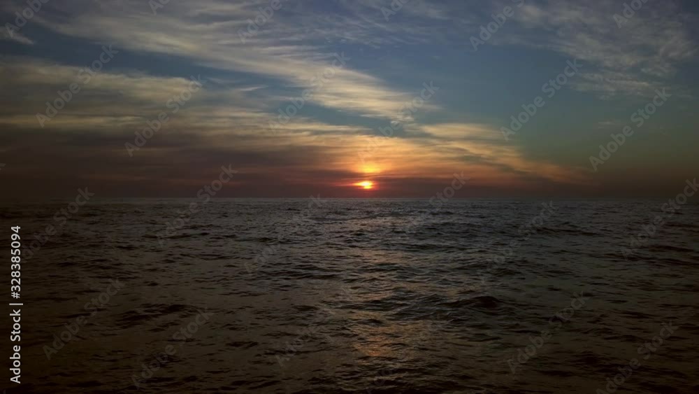 low altitude drone flight over ocean waters towards a warm orange shining sunset. Colorful gloomy sunbeam behind vibrant colorful glowing clouds. Scenic panoramic wide angle with mystical summer dusk
