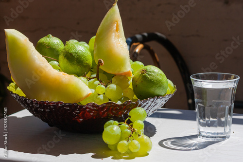 Glass of water and plate with fruit - grapes, lemons, melon slices on the table in a sunny day. photo