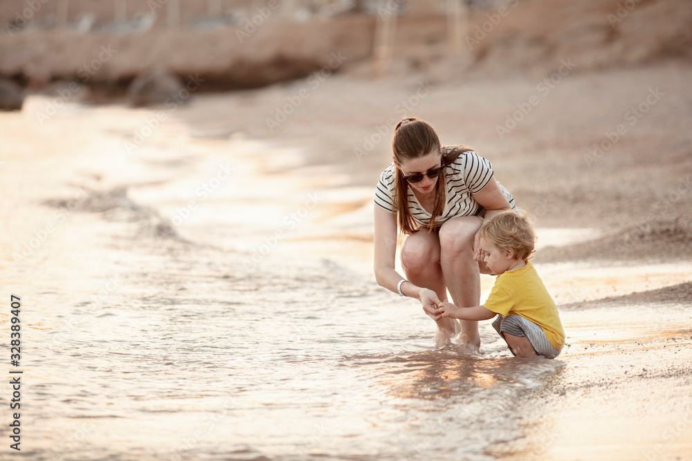 Happy mother and toddler play on the seaside. Mom and baby play in the sea beach. Mother's love. Lifestyle. Summer rest. Family vacation. Mothers day. Happy mother and toddler play on the seaside. Mom and baby play in the sea beach. Mother's love. Lifestyle. Summer rest. Family vacation. Mothers day.