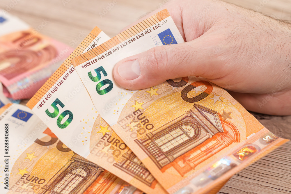 Męska dłoń trzyma dwa banknoty o nominale 50 Euro. Na drugim planie banknoty euro leżą na stole. - obrazy, fototapety, plakaty 