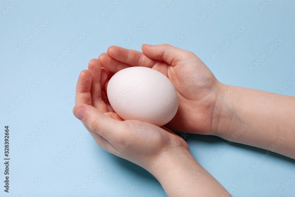 White chicken egg in the hands of a child on a blue background. Easter concept. White chicken egg in the hands of a child on a blue background. Easter concept.
