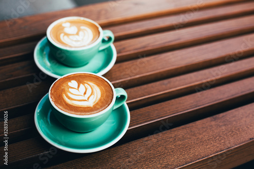 Two cups of cappuccino with latte art on wooden background. Beautiful foam, greenery ceramic cups, stylish toning, place for text.
