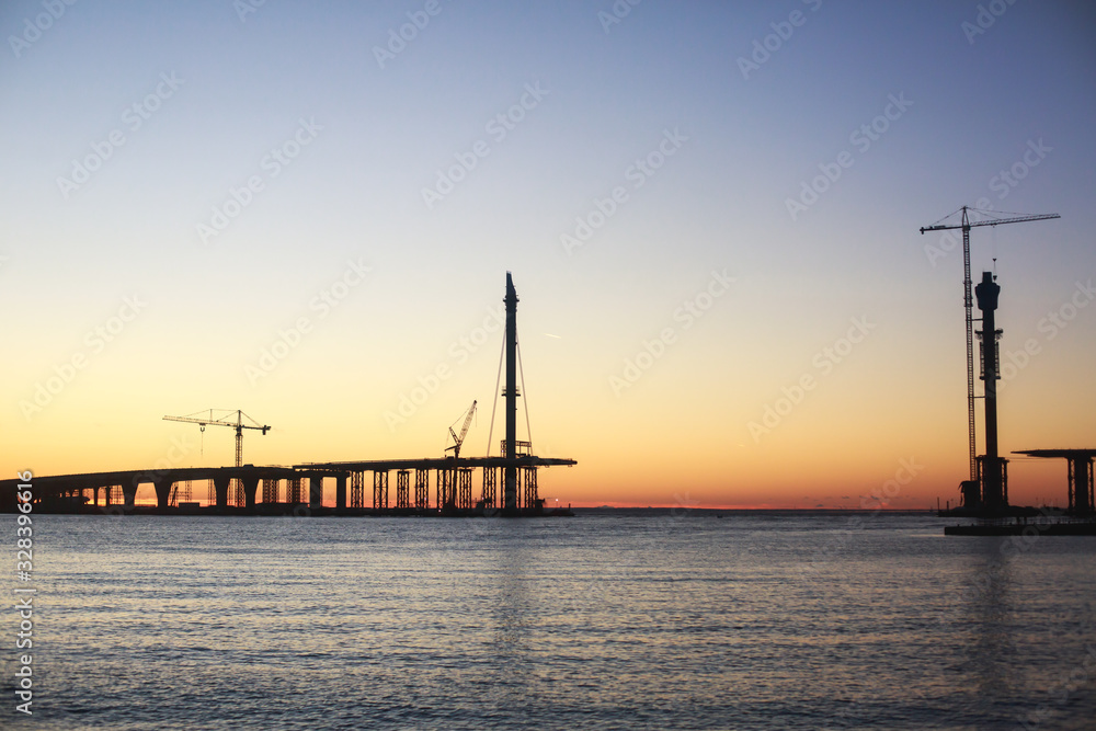An unfinished part of highway road or bridge, on sunset or dusk, Industrial construction cranes and building silhouettes at sunrise, back light construction site silhouetted at sunset, over the water