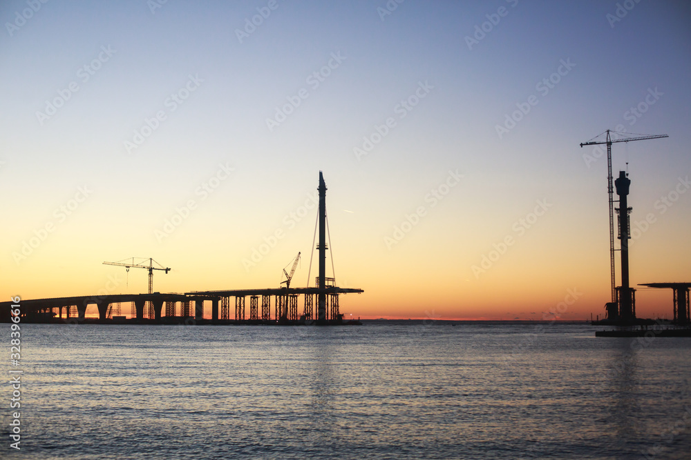 An unfinished part of highway road or bridge, on sunset or dusk, Industrial construction cranes and building silhouettes at sunrise, back light construction site silhouetted at sunset, over the water An unfinished part of highway road or bridge, on sunset or dusk, Industrial construction cranes and building silhouettes at sunrise, back light construction site silhouetted at sunset, over the water