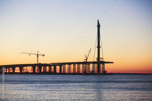 An unfinished part of highway road or bridge  on sunset or dusk  Industrial construction cranes and building silhouettes at sunrise  back light construction site silhouetted at sunset  over the water