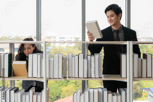 asian male and cuacasian female university students standing at bookshelf seaching and reading book in library photo