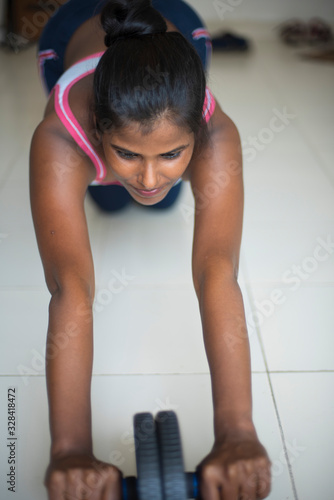 Indian dark skinned brunette girl in sportswear performing exercise with ab roller in front of glass window in a white background. Indian lifestyle