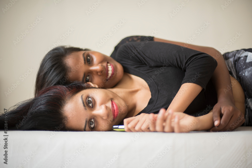 Obraz premium Two black and white attractive young brunette Indian Bengali women in black western dress are sleeping together in white bed in white studio background. Indian lifestyle.