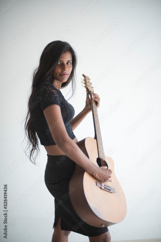 Portrait of an attractive young brunette dark skinned Indian Bengali girl in black western sportswear playing guitar in white studio background. Indian lifestyle Portrait of an attractive young brunette dark skinned Indian Bengali girl in black western sportswear playing guitar in white studio background. Indian lifestyle