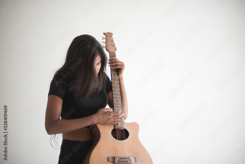 Portrait of an attractive young brunette dark skinned Indian Bengali girl in black western sportswear playing guitar in white studio background. Indian lifestyle Portrait of an attractive young brunette dark skinned Indian Bengali girl in black western sportswear playing guitar in white studio background. Indian lifestyle