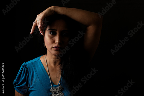 Close up face portrait of a beautiful Indian Bengali brunette woman in light and shadow before a black copy space background wearing a blue top. Indian lifestyle and fashion portrait
