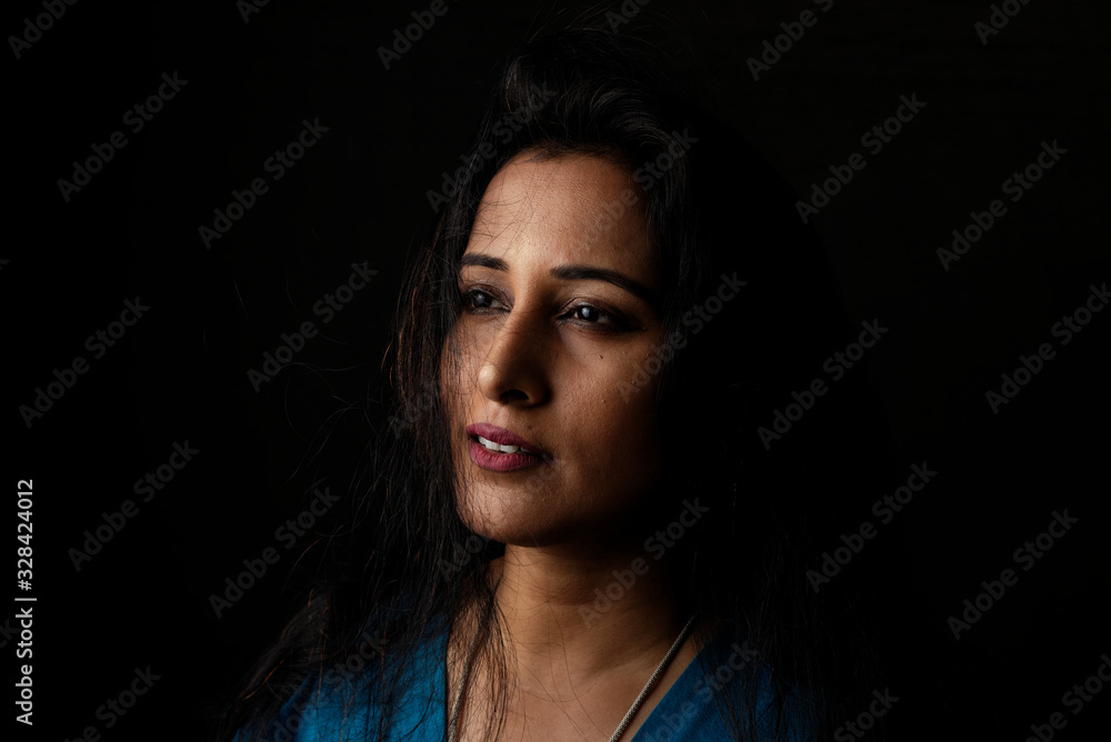 Close up face portrait of a beautiful Indian Bengali brunette woman in light and shadow before a black copy space background wearing a blue top. Indian lifestyle and fashion portrait Close up face portrait of a beautiful Indian Bengali brunette woman in light and shadow before a black copy space background wearing a blue top. Indian lifestyle and fashion portrait