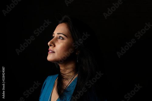 Close up face portrait of a beautiful Indian Bengali brunette woman in light and shadow before a black copy space background wearing a blue top. Indian lifestyle and fashion  portrait